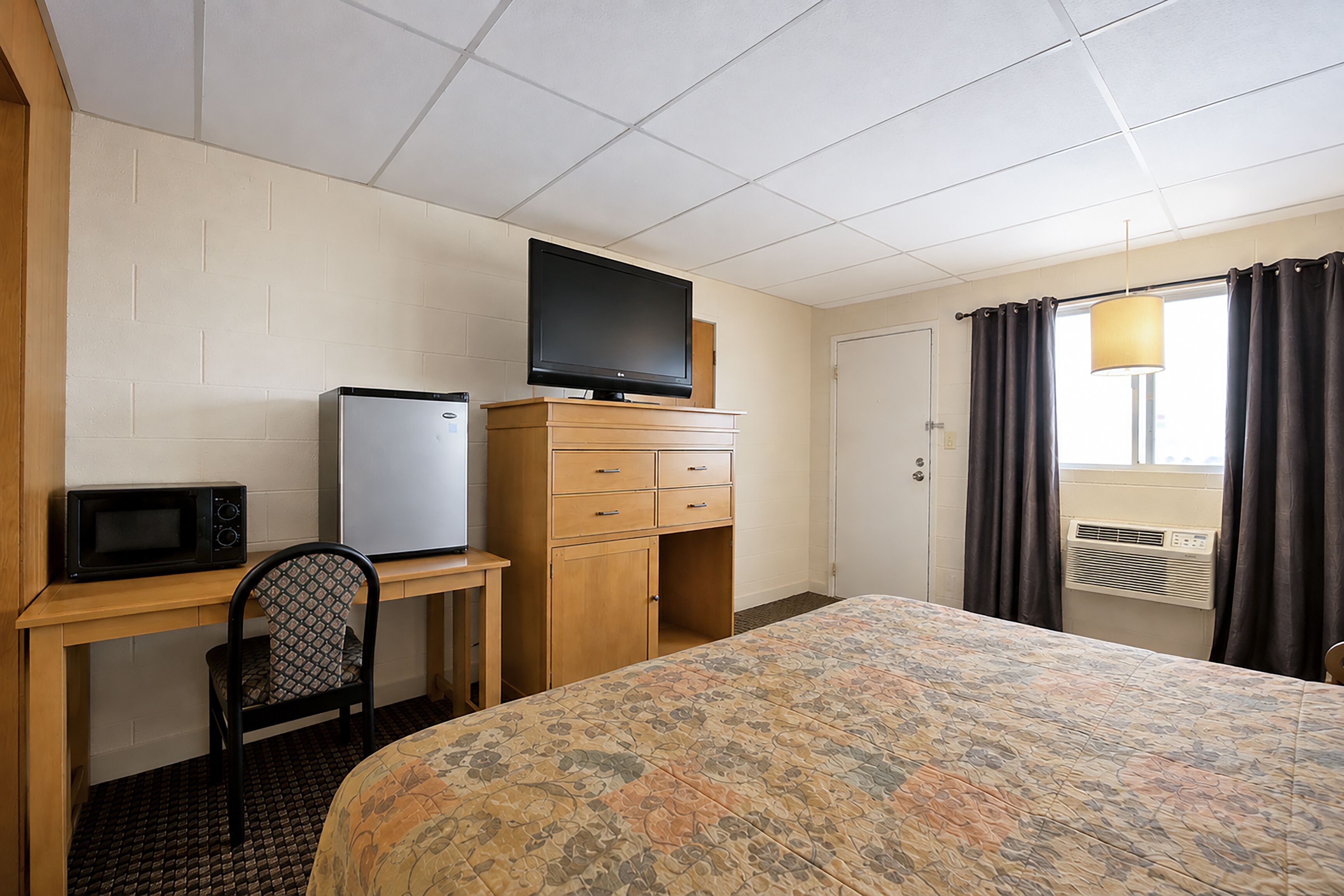 A hotel room with a bed, a desk with a computer, a television, a refrigerator, and a chair.