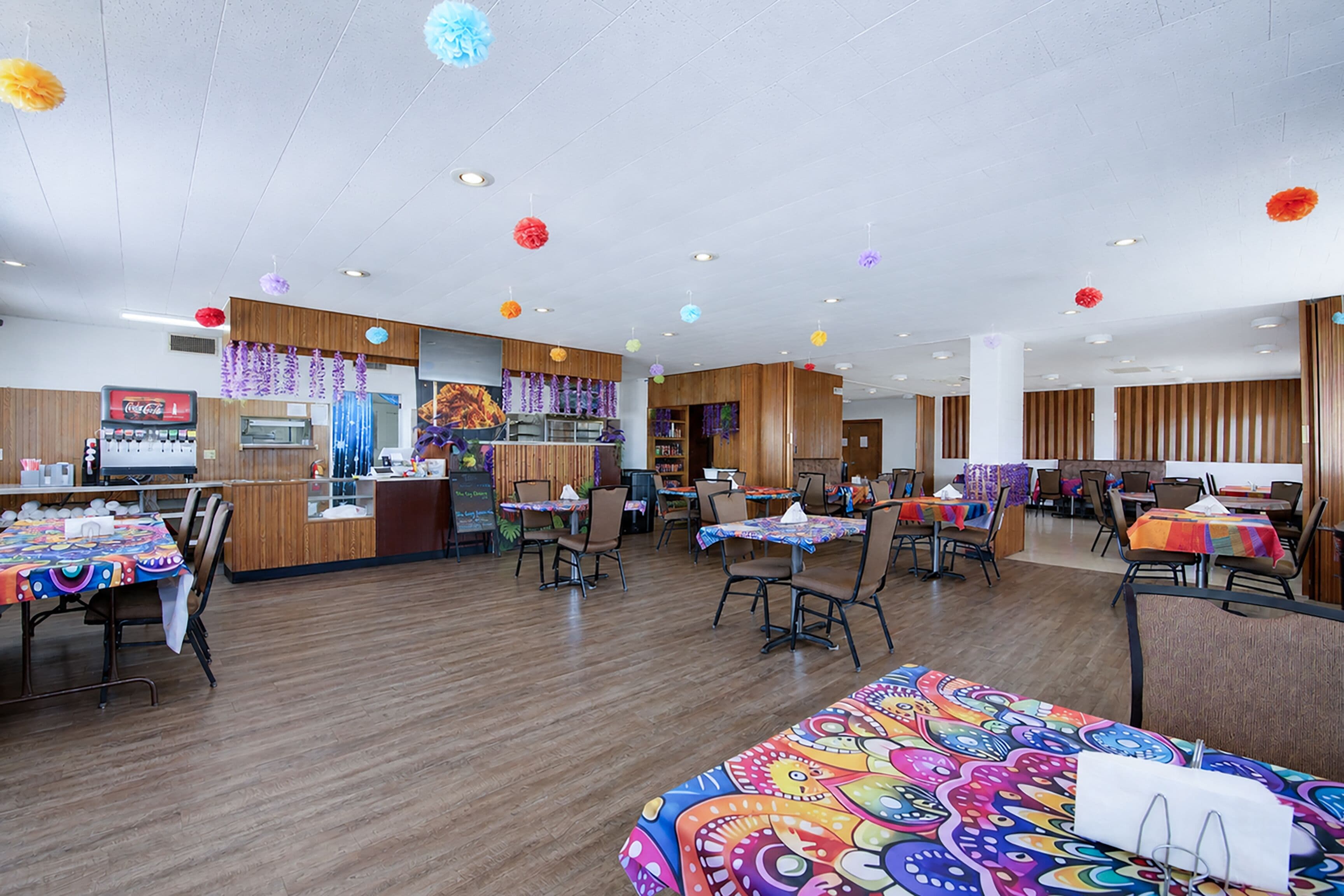 Um restaurante com piso de madeira, mesas com toalhas coloridas e decorações penduradas.