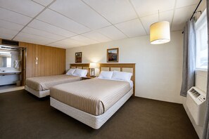 A hotel room with two beds, a wooden headboard, a window with curtains, a wall-mounted heater, and a small table with a lamp. - First Inn By OYO Quinter Hwy 70 (Quinter)