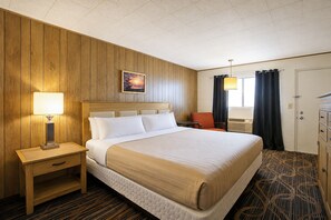 A hotel room with a wooden paneled wall, a bed with white linens, a nightstand with a lamp, a painting on the wall, and a window with curtains. - First Inn By OYO Quinter Hwy 70 (Quinter)