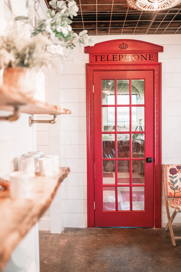 A red telephone booth with a crown emblem, a wooden shelf with jars, and a wicker chair.
