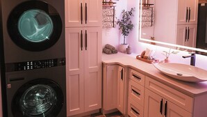 A compact laundry room with two stacked front-loading washing machines, a countertop with a sink and mirror, and a hexagonal tile floor. - Pickleball Cafe & Micro Hotel (Ironton)