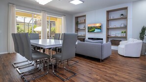 A modern living room with a dining area, a grey sofa set, a TV mounted on the wall, and wooden shelves with decorative items. - Pristine Fully Renovated House - 12min from Disney Parks (Kissimmee)