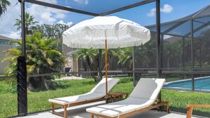 A glass-enclosed lounge area with two white lounge chairs and a white umbrella, overlooking a pool and lush greenery. - Pristine Fully Renovated House - 12min from Disney Parks (Kissimmee)