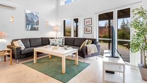 Un salón amplio con un sofá modular grande, una mesa de centro y un televisor. Hay ventanas grandes que ofrecen luz natural y vistas al exterior.