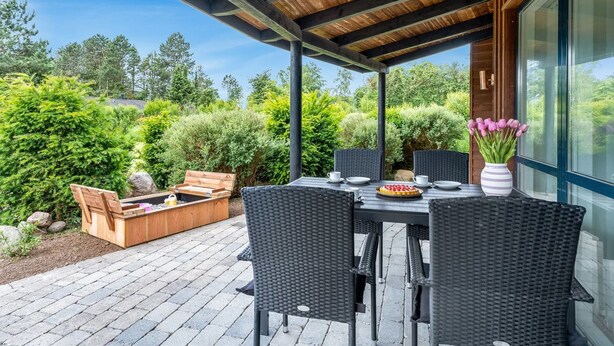 Un patio al aire libre con una pérgola de madera, sillas de mimbre, una mesa de comedor con platos y un jarrón de flores, y una chimenea.