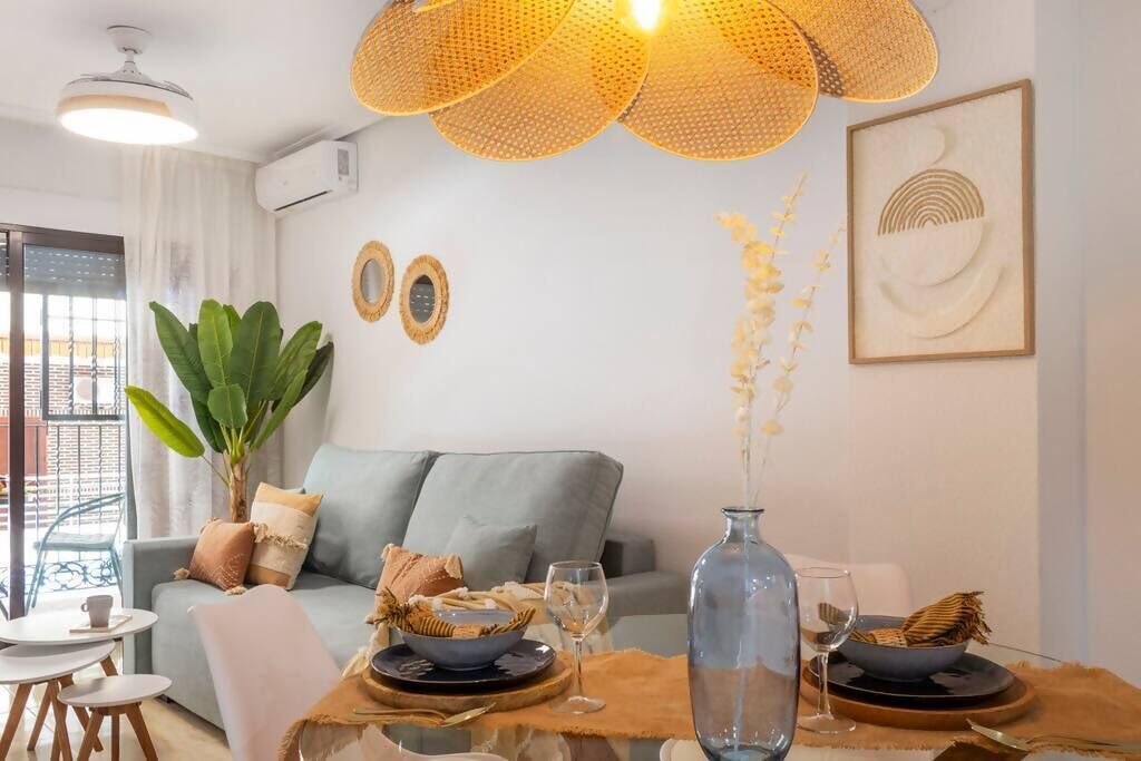 A modern living room with a grey sofa, a dining table set with plates, glasses, and a vase. There is a large plant and a hanging light fixture above the table.