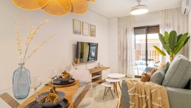 A modern living room with a glass dining table, a sofa, a TV, and a large window with curtains. - Beautiful apartment on the beach (Torrevieja)