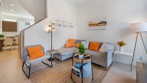 Ett modernt vardagsrum med en grå soffa, orange kuddar, ett litet sidobord, en golvlampa och en trappa som leder till ett annat rum.