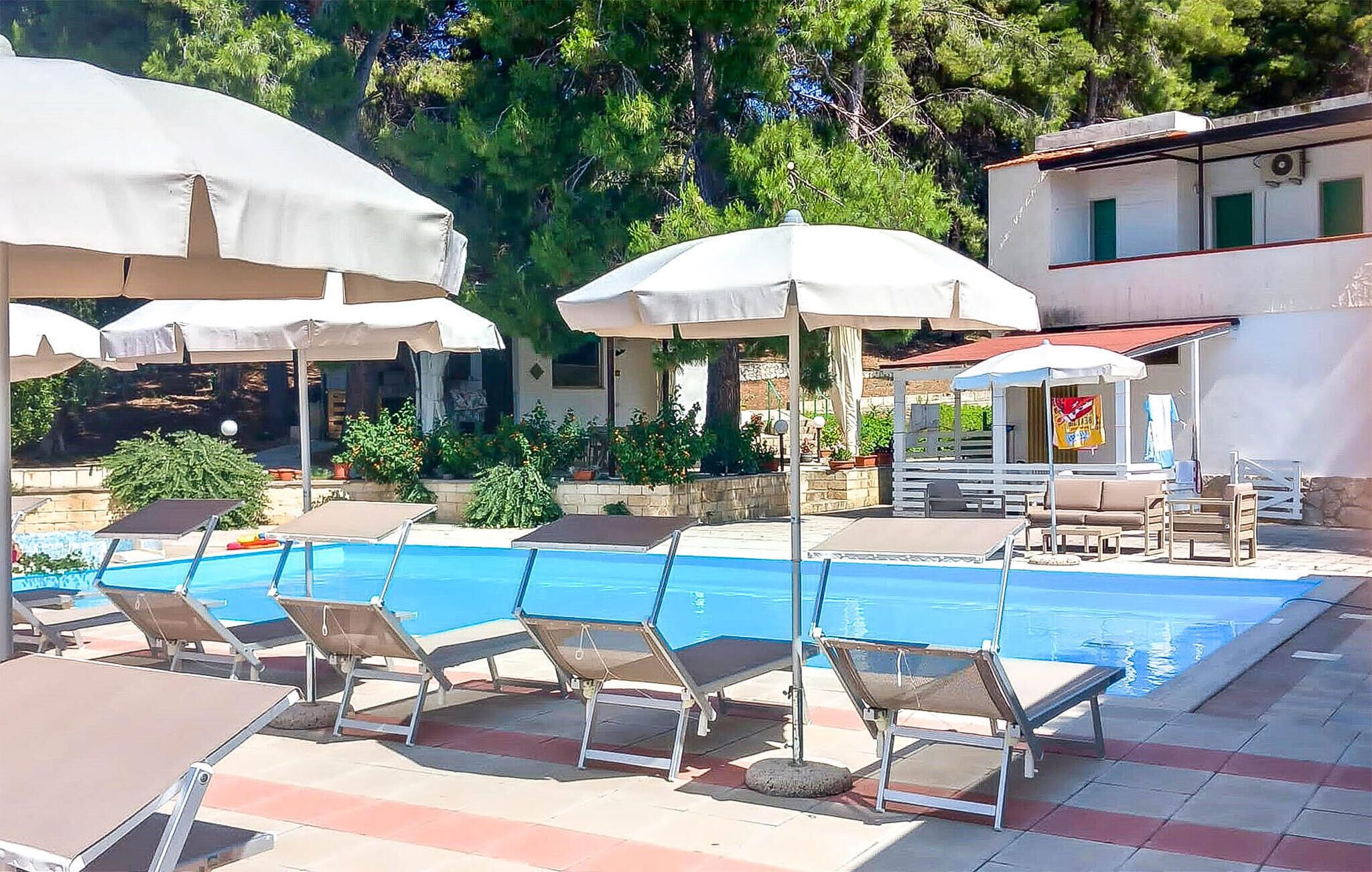 Ein Poolbereich mit Liegestühlen und Sonnenschirmen, umgeben von Grünflächen und einem Gebäude mit rotem Dach.
