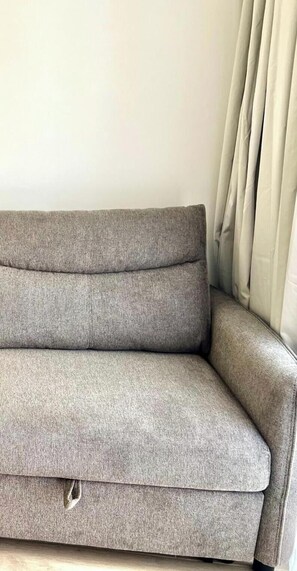 A grey sofa with a cushion, a small storage compartment, and a curtain.