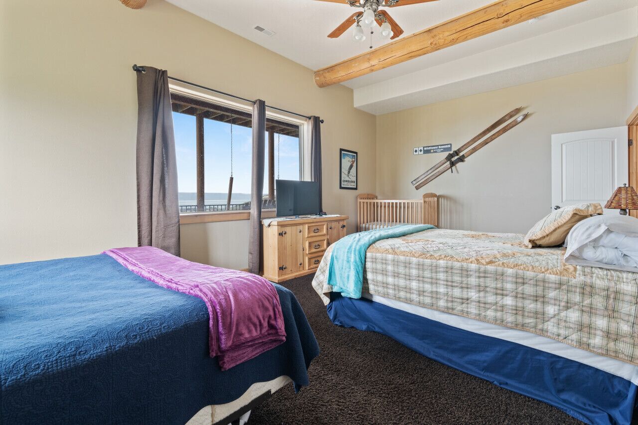 A bedroom with two beds, a wooden ceiling fan, a window with curtains, a desk with a chair, and a framed picture on the wall.