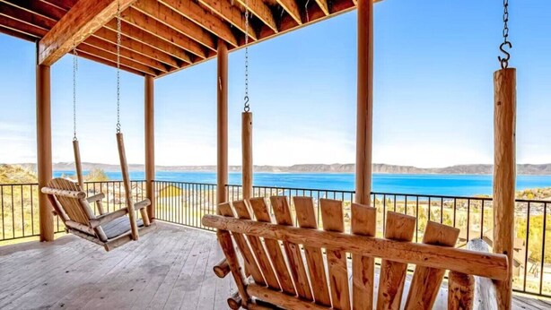 Un conjunto de columpios de madera sobre una terraza de madera, con vistas a un cuerpo de agua y colinas al fondo.