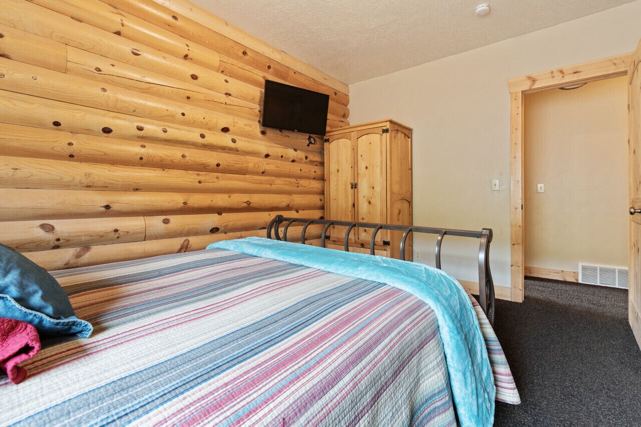 Ein Zimmer mit Wand aus Holzstämmen, einem Bett mit gestreifter Decke, einem an der Wand montierten Fernseher und einem hölzernen Kleiderschrank.
