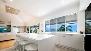 A modern kitchen with a large island, bar stools, and a view of palm trees. - Seascape on Shutehaven (Shute Harbour)