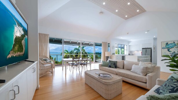 A spacious living area with a large TV, a sofa, a dining table, and a balcony with a view of the ocean. - Seascape on Shutehaven (Shute Harbour)