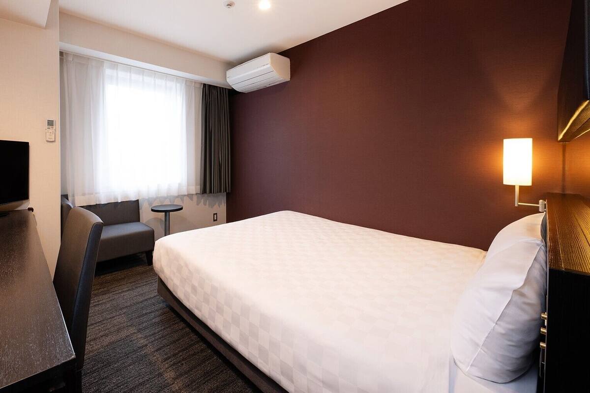 A hotel room with a bed, a desk, a chair, a TV, a window with curtains, an air conditioner, and a bedside lamp.