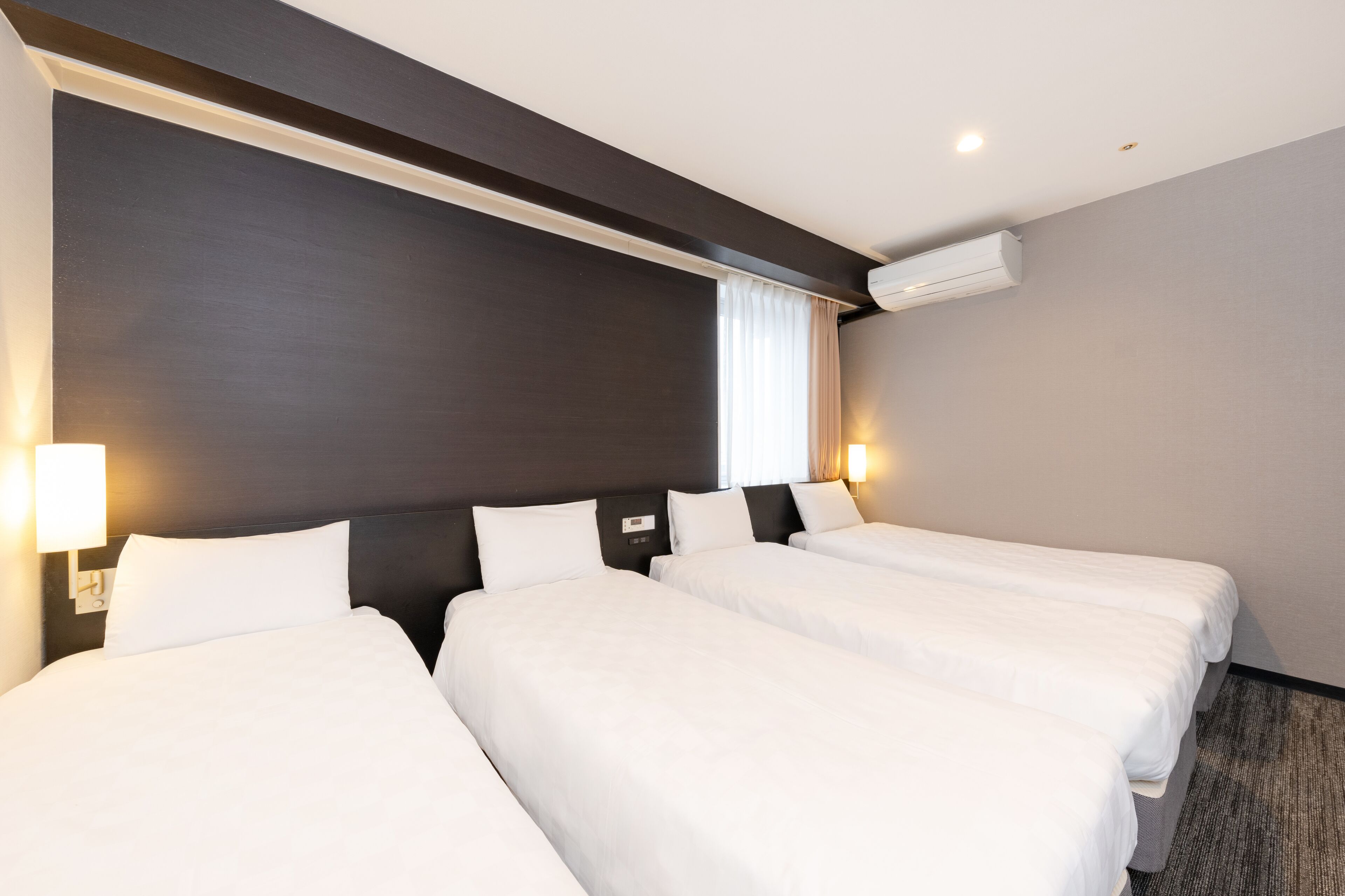 Une chambre d’hôtel avec trois lits simples, une tête de lit sombre, un climatiseur fixé au mur et deux lampes de chevet.