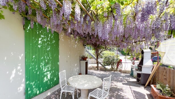 A patio with a round table and four chairs, a green door, and purple flowering vines overhead. - CHEZ CHOUCAS - LA MAISON DE CHOUCAS (Portoferraio)