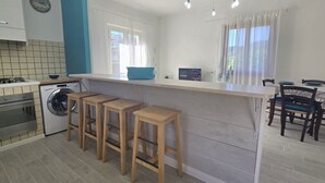 A modern kitchen with a built-in washing machine, wooden stools, a bar counter, and a dining area with chairs. - CHEZ CHOUCAS - LA MAISON DE CHOUCAS (Portoferraio)