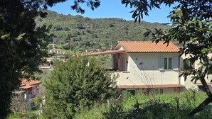 A rural house with a red-tiled roof, surrounded by dense greenery and hills in the background.