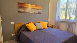 A bedroom with a bed, a nightstand, a lamp, a painting of a sunset, and a window with curtains.