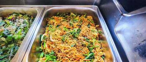 Un bufé de acero inoxidable con diversos platos de fideos, incluyendo uno con vegetales y hierbas.