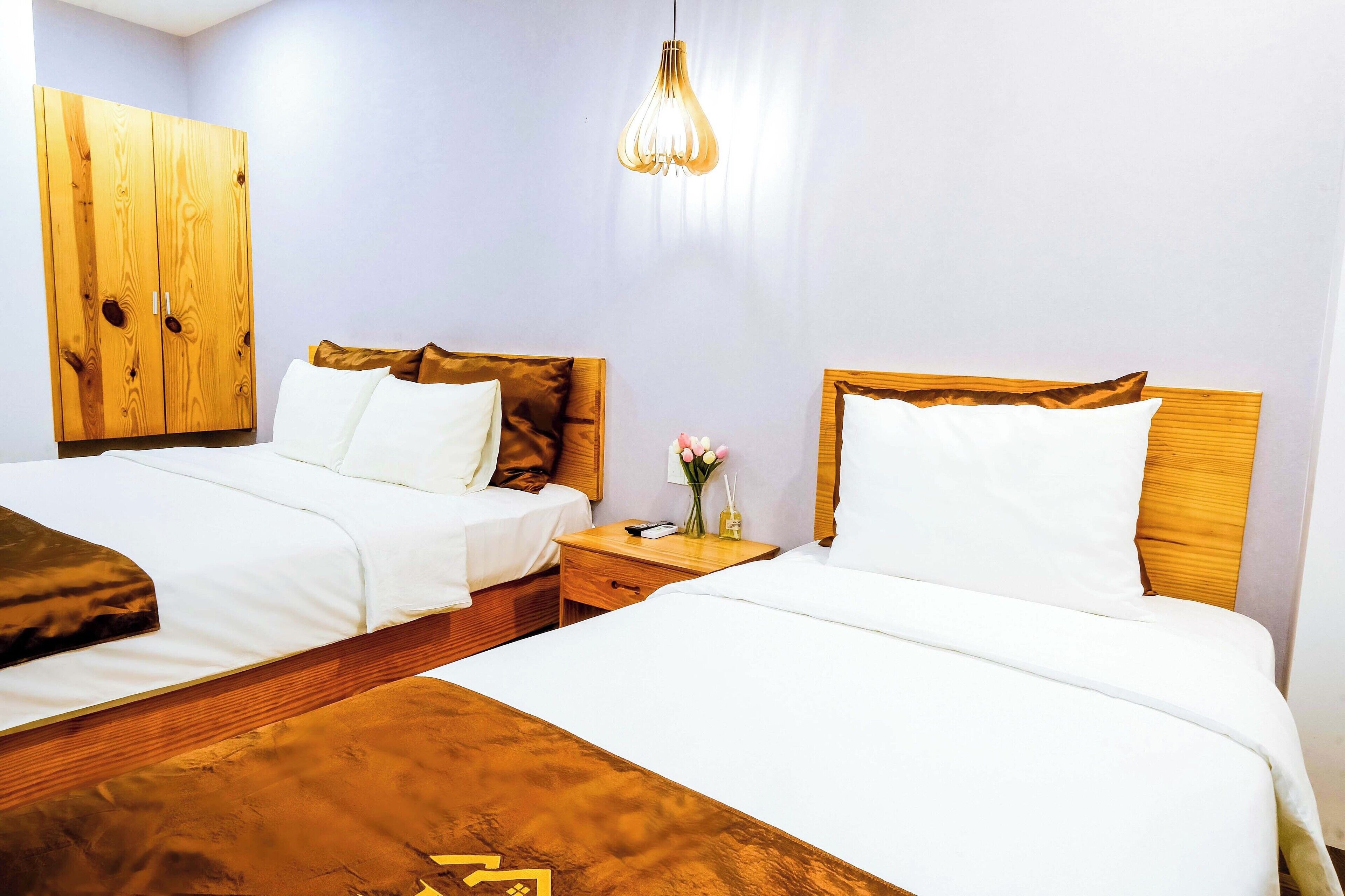 A hotel room with two beds, wooden headboards, white bedding, a nightstand with a vase of flowers, and a hanging light fixture.
