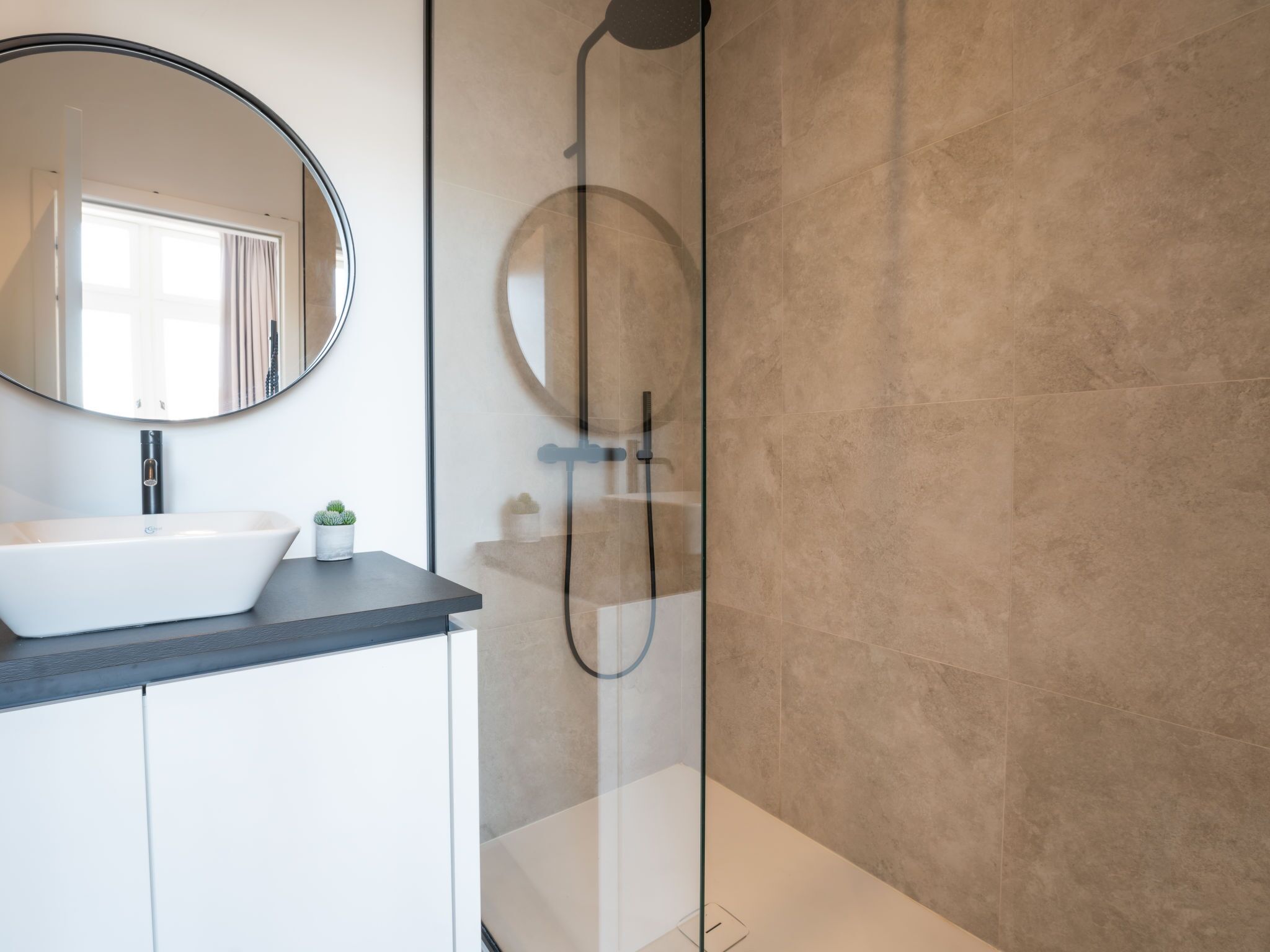 Een moderne badkamer met een wit wastafel, een ronde spiegel en een douchecabine met glazen wanden.