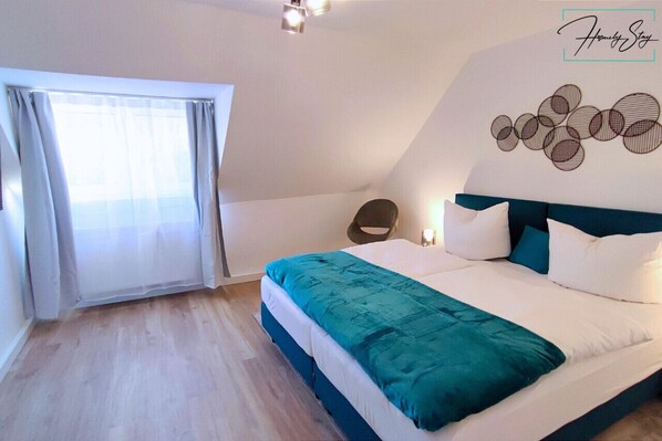 Un dormitorio con cama, una silla, una obra de arte en la pared y una ventana con cortinas.