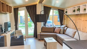 Ein modernes Wohnzimmer mit einer Couch, einem Couchtisch, einem Fernseher und einem Schreibtisch. Regale mit Pflanzen und Blick ins Freie.