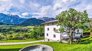 Un bain à remous dans un cadre verdoyant luxuriant, avec une montagne en arrière-plan et un bâtiment blanc doté d’un balcon.