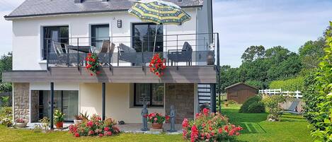 A two-story house with a balcony, a garden with flowers, and a white fence in the background.