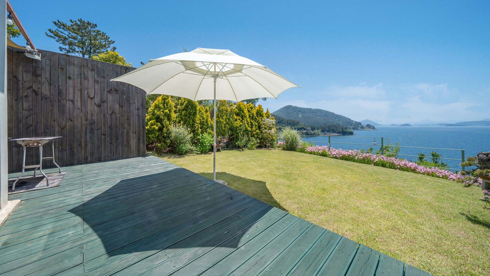 A deck with a table and an umbrella, overlooking a grassy area with flowers and a view of the sea.