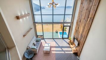 A high-ceilinged room with a large window overlooking a pool and a beach.