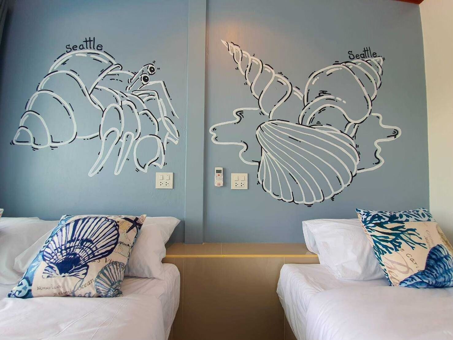 A hotel room with two beds, each with a white mattress and a pillow featuring a blue and white design. The walls are painted light blue, and there are two wall art pieces depicting stylized fish with the word "Seattle" on them.