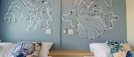 A hotel room with two beds, each with a white mattress and a pillow featuring a blue and white design. The walls are painted light blue, and there are two wall art pieces depicting stylized fish with the word "Seattle" on them.