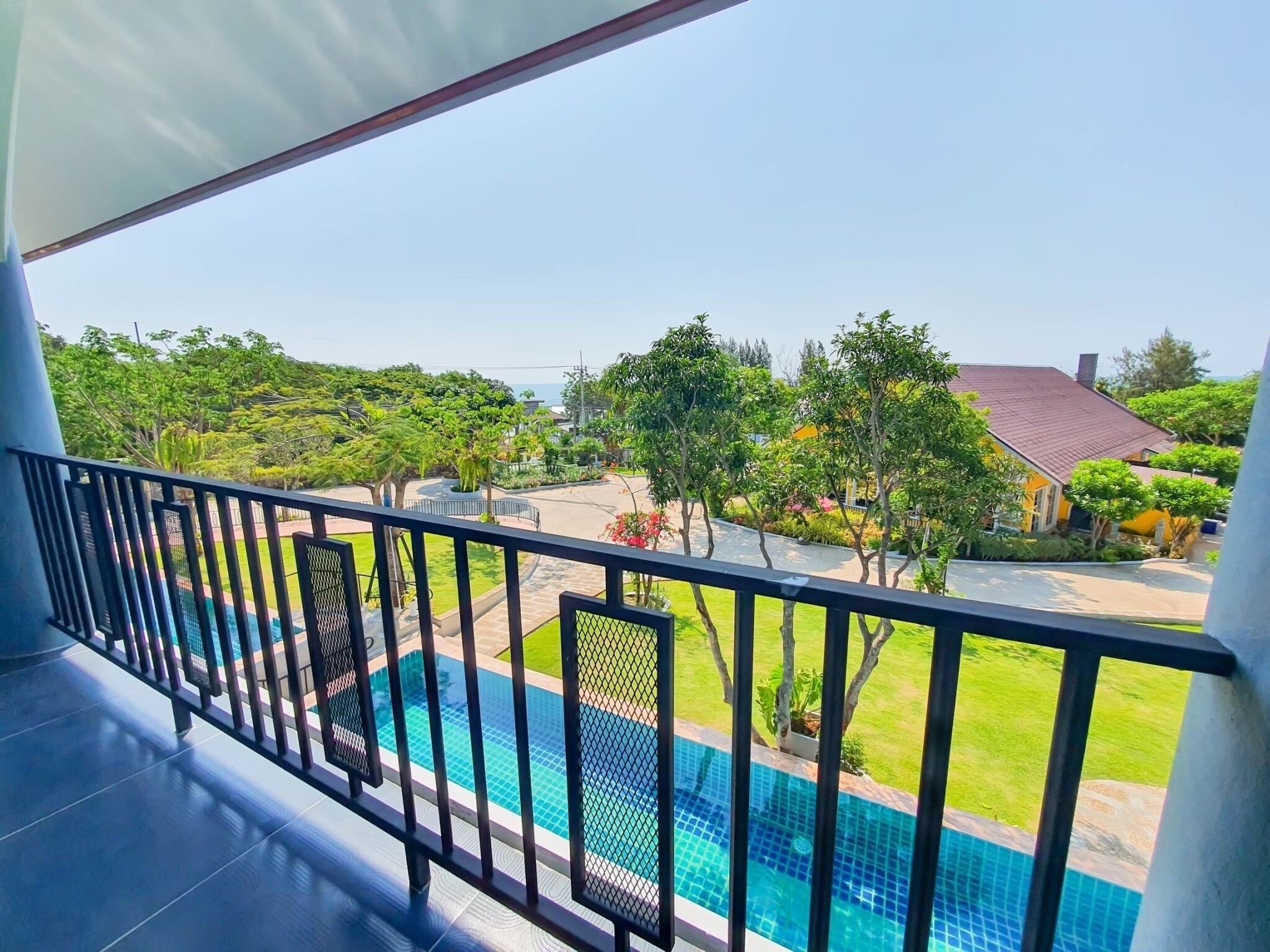 A balcony with a view of a swimming pool, lush greenery, and a building with a red roof.