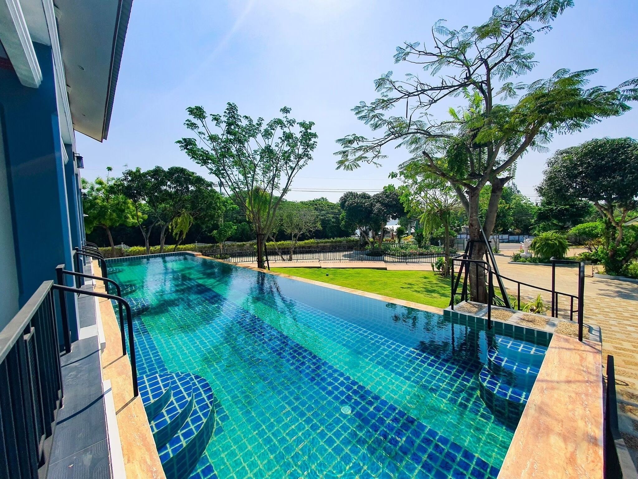 Uma piscina infinita retangular com água azul cristalina, rodeada por um deck de madeira e um guarda-corpo de metal.