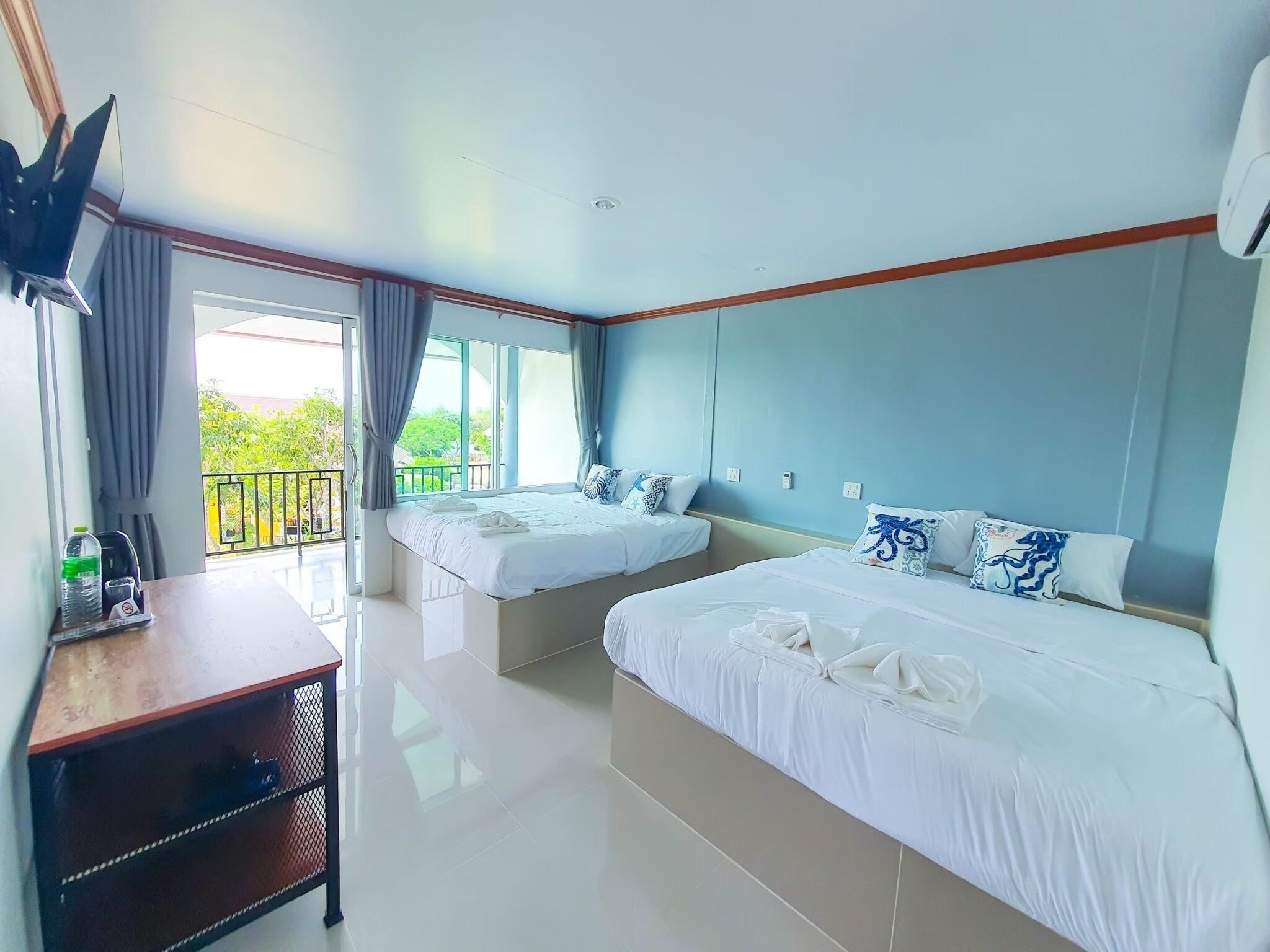 A hotel room with two beds, a TV, a desk, and a balcony with curtains.