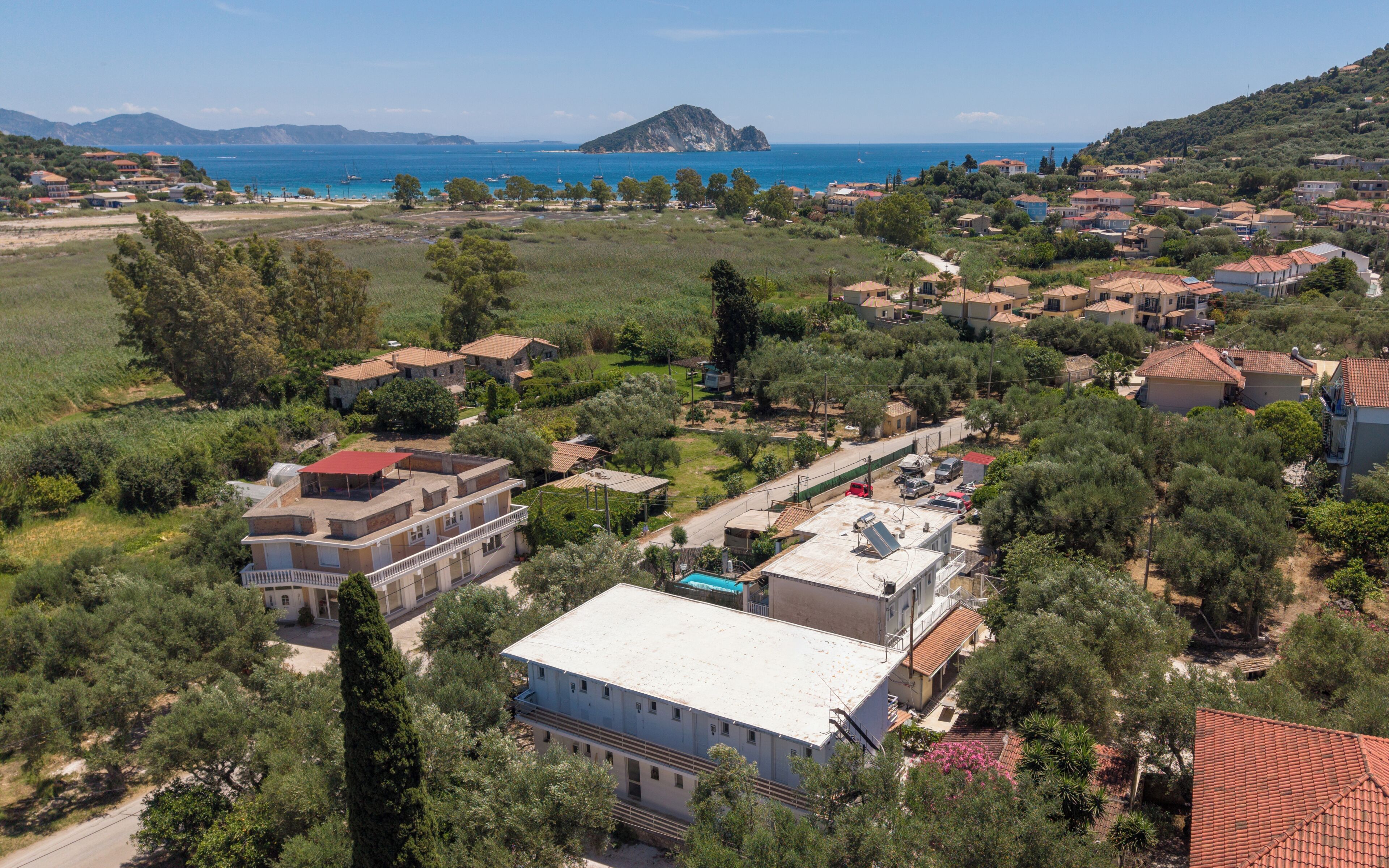 A coastal residential area with houses, a large building with a white facade, and a view of the sea and a distant island.