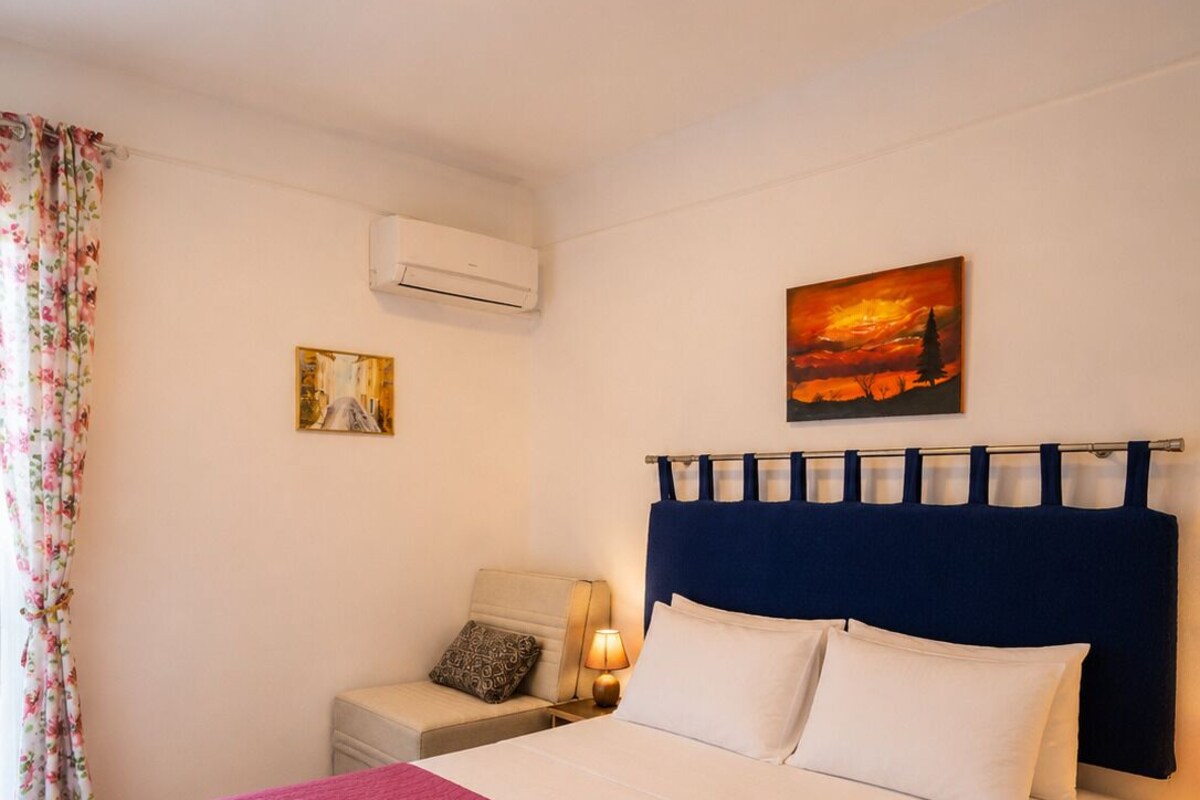 A bedroom with a bed, a nightstand, a lamp, an air conditioning unit, and a painting on the wall.