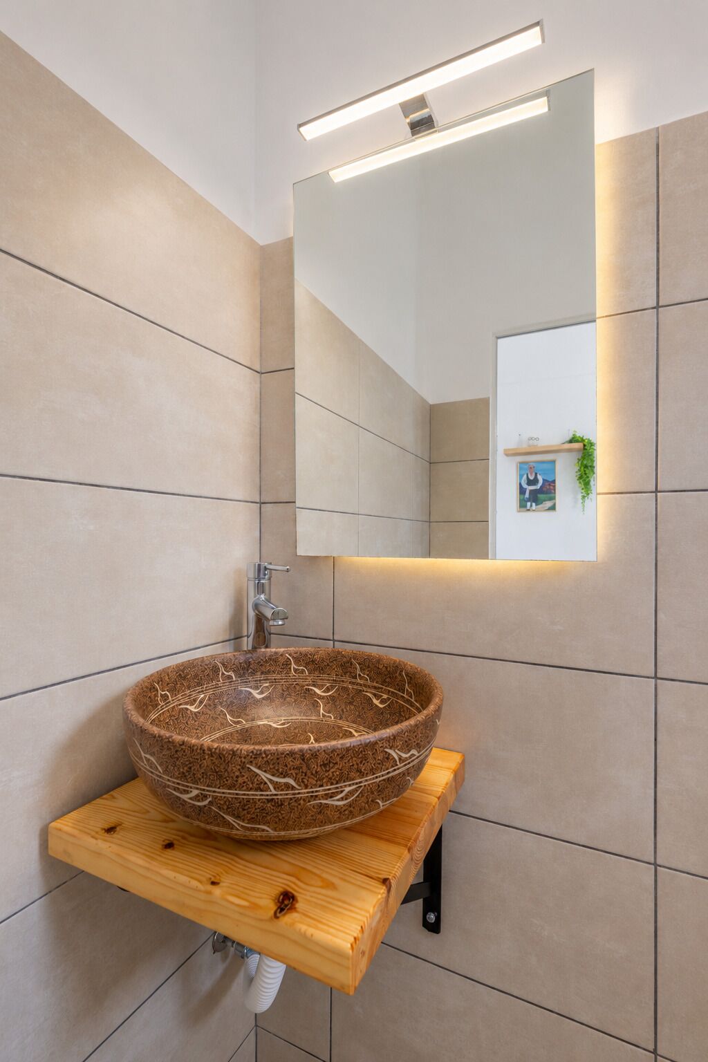 A modern bathroom with a large vessel sink on a wooden shelf, a wall-mounted mirror, and a small shelf with a plant.