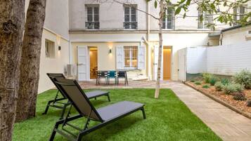 Un patio con césped artificial, dos sillones, una mesa con sillas y un pequeño árbol.