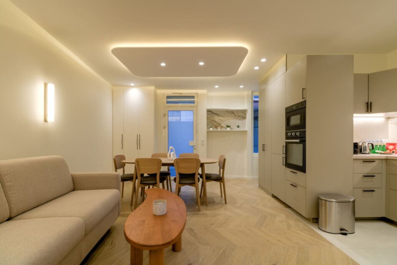 Una sala moderna con un sofá, una mesa de centro y un comedor con sillas y una mesa. Hay una cocina con armarios, un microondas y un cubo de basura.