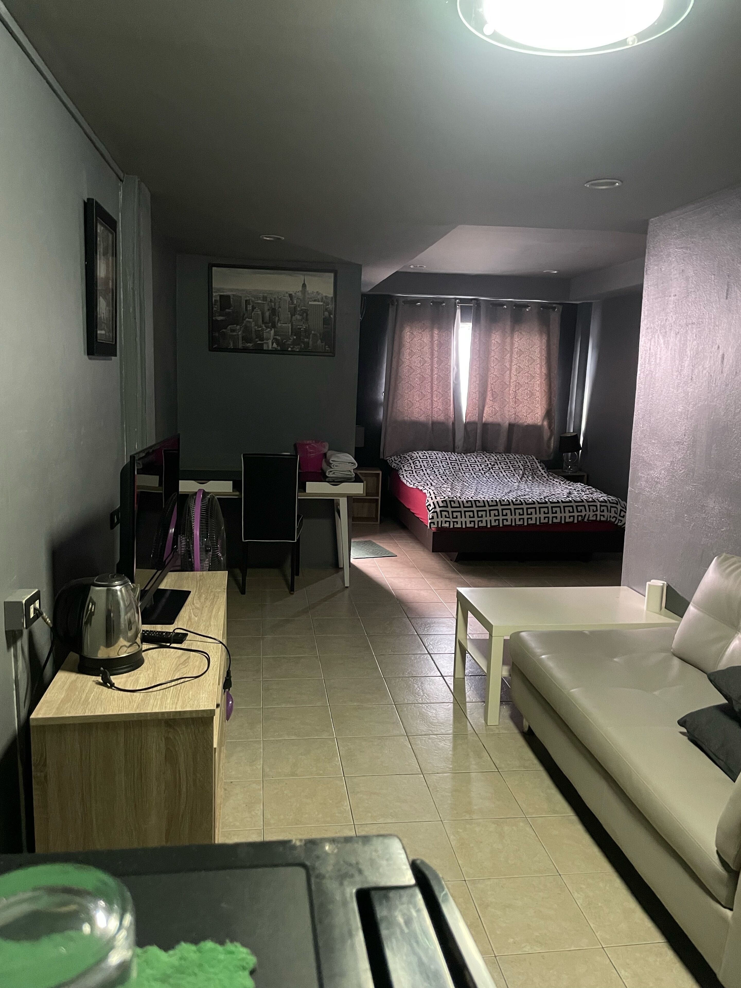 A compact hotel room with a bed, a sofa, a desk with a chair, a small table, and a kettle.