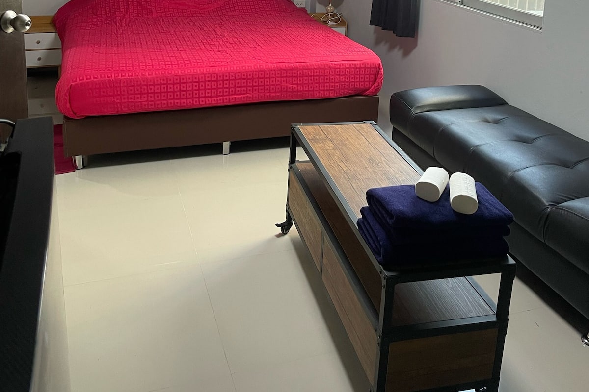 A bedroom with a large bed, a bedside table, a bench, a window, and an air conditioning unit.