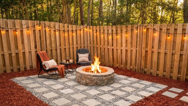 A backyard with a stone fire pit, two chairs, a table, and string lights. - Quiet, Pet Friendly Townhome - 2 Kings/1 Queen, Fire Pit, close to downtown  (Greensboro)