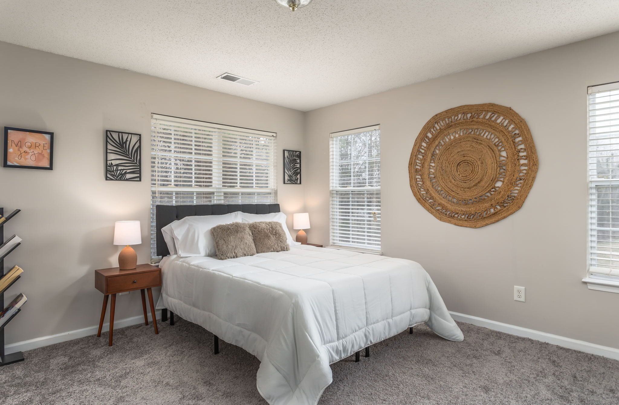 A bedroom with a bed, two bedside tables, a wall-mounted circular rug, and a window with blinds.