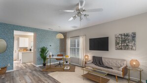 A living room with a sofa, coffee table, and dining area. There is a ceiling fan, a TV mounted on the wall, and a window with curtains. - Quiet, Pet Friendly Townhome - 2 Kings/1 Queen, Fire Pit, close to downtown  (Greensboro)